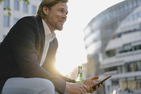 Geschäftsmann sitzt auf einer Bank in der Stadt bei Sonnenuntergang mit einer Flasche Bier und Smartphone, lizenzfreies Stockfoto