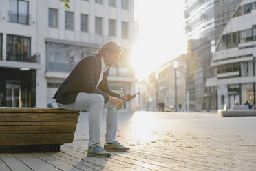 Geschäftsmann sitzt auf einer Bank in der Stadt bei Sonnenuntergang mit einer Flasche Bier und Smartphone - JOSEF00921