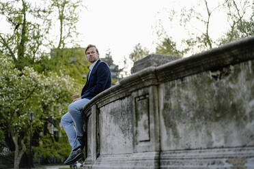 Relaxed businessman with takeaway coffee sitting on a wall in city park - JOSEF00936