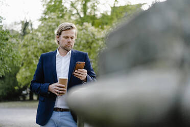 Businessman with takeaway coffee using smartphone in city park - JOSEF00937