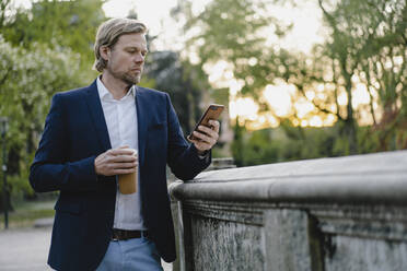 Businessman with takeaway coffee using smartphone in city park - JOSEF00938