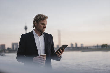 Businessman with takeaway coffee and tablet at the riverside at dusk - JOSEF00944