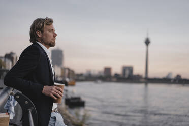 Businessman with takeaway coffee at the riverside at dusk - JOSEF00947