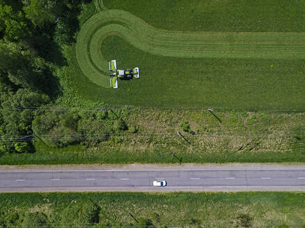 Russia, Moscow Oblast, Aerial view of car driving along country road past tractor mowing green field - KNTF04711