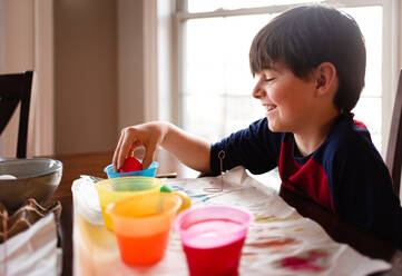Ein fröhlicher Junge taucht ein Ei in eine Schale mit Farbstoff, um es für Ostern zu färben. - CAVF85564