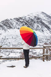 Frau mit Regenbogenschirm vor einer verschneiten Berglandschaft - CAVF85573