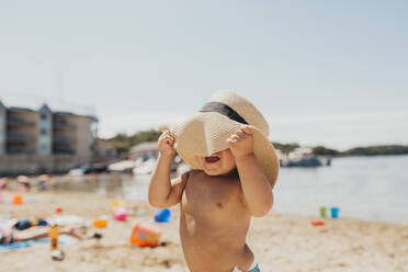 Toddler Boy Playing Peek-a-boo with Sun Hat on Beach - CAVF85624