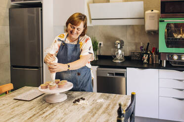 Lächelnde reife Frau, die Cupcakes auf einem Tortenständer in einer Werkstatt glasiert - GMLF00235