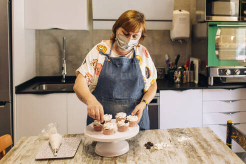 Bäckerin mit Maske beim Verzieren von Törtchen auf einem Kuchenstand in einer Werkstatt - GMLF00237