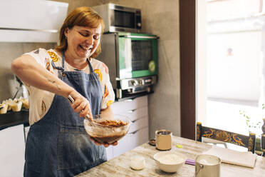 Lächelnde Bäckerin mischt Kuchenteig in einer Schüssel auf einem Tisch in einer Werkstatt - GMLF00238
