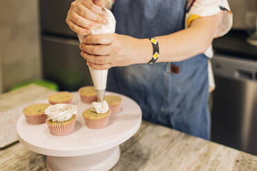 Nahaufnahme einer reifen Frau, die in einer Werkstatt Cupcakes auf einem Tortenständer glasiert - GMLF00239