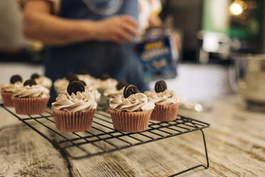 Nahaufnahme von Cupcakes auf einem Gestell mit einer Bäckerin im Hintergrund in einem Geschäft - GMLF00241