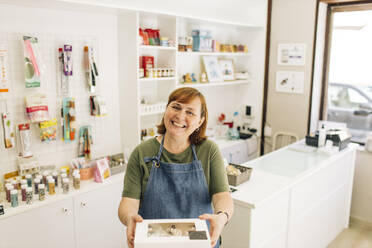 Smiling female baker delivering cupcake box while standing in store - GMLF00244