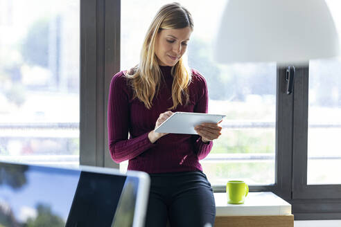 Geschäftsfrau, die ein digitales Tablet benutzt, während sie im Büro am Fenster sitzt - JSRF00960