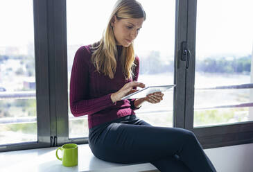 Geschäftsfrau, die ein digitales Tablet benutzt, während sie auf dem Tisch vor dem Fenster im Büro sitzt - JSRF00961