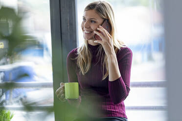 Lächelnde Unternehmerin im Gespräch über ein Smartphone, während sie im Büro am Fenster sitzt - JSRF00963