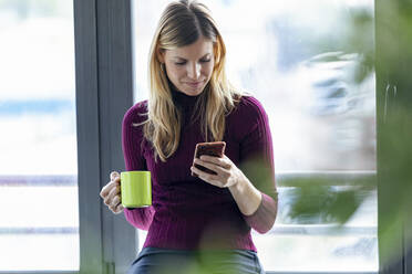 Businesswoman holding coffee mug using smart phone while sitting in office - JSRF00964