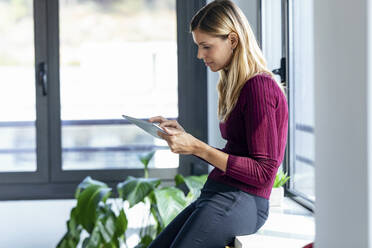 Eine Unternehmerin benutzt ein digitales Tablet, während sie sich im Büro auf einen Tisch stützt - JSRF00965