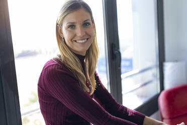 Lächelnde Geschäftsfrau, die im Büro am Fenster sitzt - JSRF00968