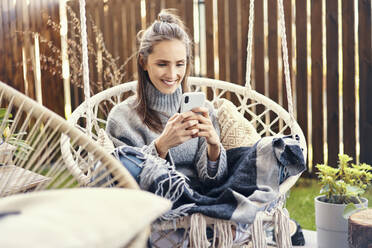 Lächelnde junge Frau benutzt ihr Smartphone, während sie sich auf einer Schaukel im Garten entspannt - BSZF01560