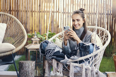 Lächelnde schöne Frau, die ein Smartphone benutzt, während sie sich auf einer Schaukel im Garten entspannt - BSZF01561