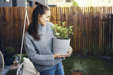 Glückliche junge Frau, die eine Topfpflanze trägt, während sie im Garten steht - BSZF01569