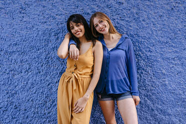 Smiling female friends standing against blue wall in city - TCEF00761