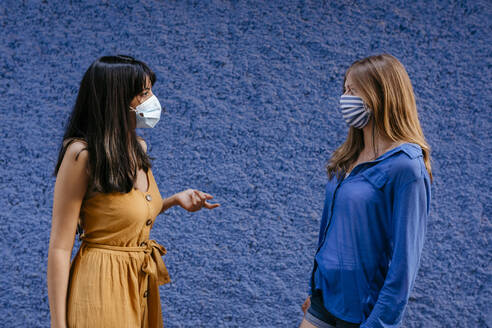 Female friends wearing masks talking while maintaining distance against blue wall in city - TCEF00762