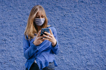 Young woman wearing mask using smart phone while standing against wall in city - TCEF00764