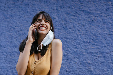 Nahaufnahme einer lächelnden Frau mit Maske, die über ein Smartphone an einer Wand in der Stadt spricht - TCEF00765