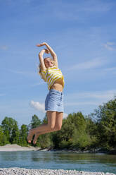 Carefree young woman with arms raised jumping at riverbank against sky - LBF03077