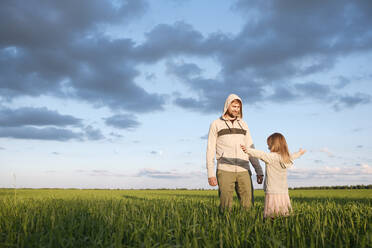 Mann schaut auf seine Tochter, die mit ausgestreckten Armen auf einer Graslandschaft steht - EYAF01155