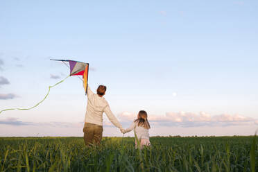 Man holding kite while walking with daughter amidst plants on green landscape - EYAF01161