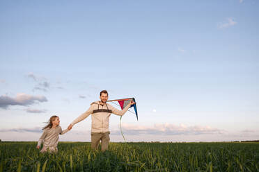 Fröhlicher Vater und Tochter mit Drachen auf grüner Landschaft gegen den Himmel laufend - EYAF01163