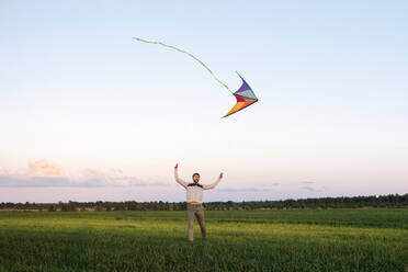 Mid adult man flying kite while standing on grassy landscape against sky at sunset - EYAF01166