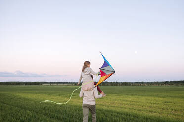 Vater, der seine Tochter auf den Schultern trägt, während er bei Sonnenuntergang über eine Graslandschaft läuft - EYAF01167