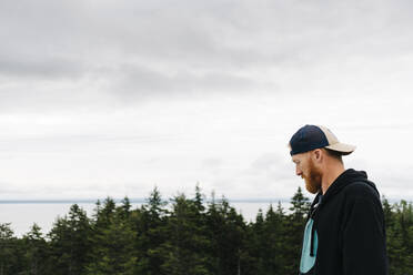 Bearded handsome man wearing cap at Bay of Fundy against sky - JVSF00016