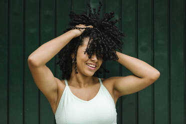 Happy young woman with hand in black curls while standing against green metal door - XLGF00227
