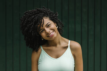 Smiling young beautiful woman with black curly hair against green metal door - XLGF00230