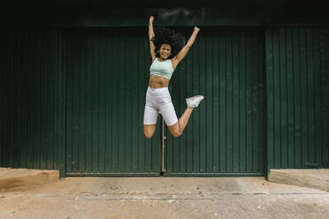 Aufgeregte junge Frau springt gegen grüne Metalltür - XLGF00231