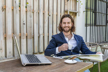 Lächelnder gutaussehender junger Geschäftsmann, der Zeitung liest, während er eine Kaffeetasse neben einem Laptop am Tisch in einem Café hält - DLTSF00780