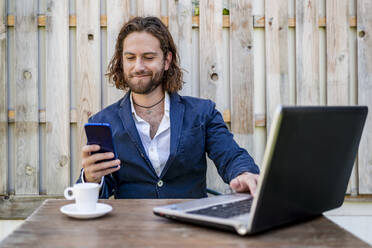 Geschäftsmann, der grinsend ein Mobiltelefon benutzt, während er in einem Café sitzt - DLTSF00783