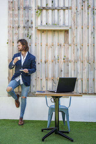 Fashionable young businessman drinking espresso by laptop on table against wooden wall at cafe stock photo