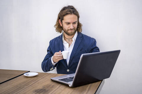 Junger männlicher Unternehmer, der Kaffee trinkt und einen Laptop an der weißen Wand eines Cafés benutzt, lizenzfreies Stockfoto