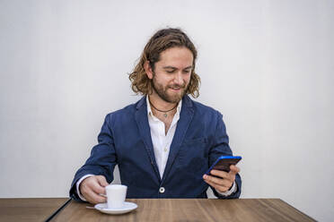 Smiling handsome businessman using smart phone while sitting with espresso at table against white wall in cafe - DLTSF00789