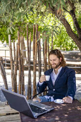 Smiling businessman drinking espresso while sitting with laptop at cafe - DLTSF00790