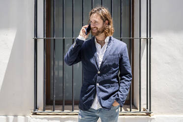 Handsome smiling male professional talking on mobile phone while standing against window - DLTSF00796
