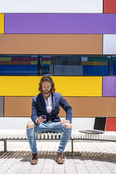 Junger männlicher Unternehmer, der sein Smartphone benutzt, während er an einem sonnigen Tag auf einer Bank vor einer bunten Wand sitzt - DLTSF00805