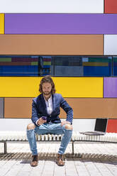 Young male entrepreneur using smart phone while sitting on bench against colorful wall during sunny day - DLTSF00805