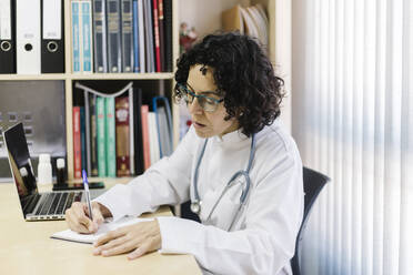 Female doctor writing by laptop while sitting at desk in office - XLGF00252